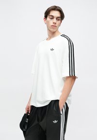 White oversized t-shirt with black three-stripe accents on sleeves and small black logo on chest; smooth cotton fabric. Black sports pants visible.