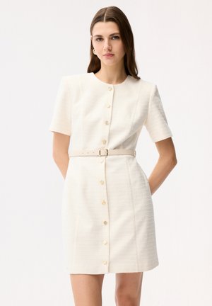 Donna che indossa un vestito corto a maniche corte color crema con bottoni dorati frontali e una cintura beige, in piedi davanti a uno sfondo bianco semplice.