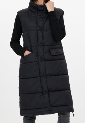 Frau trägt eine lange schwarze gesteppte ärmellose Weste mit vorderen Druckknöpfen und Seitentaschen über einem schwarzen Langarmshirt.