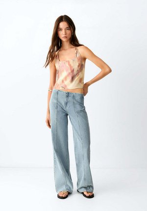 Femme aux longs cheveux bruns porte un top sans manches tie-dye, un jean taille haute à jambes larges et des sandales noires, debout contre un mur blanc uni.