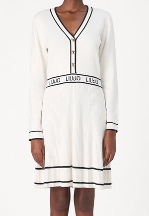 Kvinde iført en hvid plisseret kjole med lange ærmer, sort kant, guldknapper og "LIU JO"-logo ved taljen.