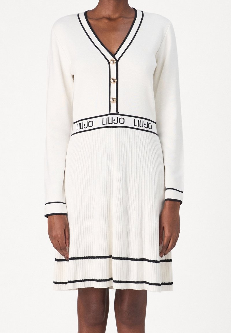 Femme portant une robe plissée blanche à manches longues, garnie de bordures noires, boutons dorés, et logo "LIU JO" à la taille.