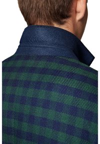 Dos d’un homme aux cheveux courts et bruns portant une veste à carreaux verte et bleu marine avec un col bleu marine relevé.