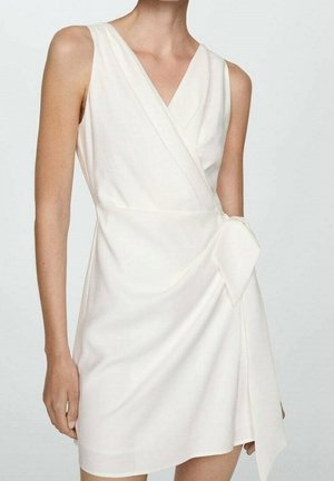 Bílé bezrukávové zavinovací šaty s výstřihem do V a mašlí na boku, nošené stojící osobou na jednobarevném světlém pozadí.