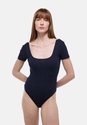 Femme aux cheveux bruns lisses portant un body côtelé bleu marine à manches courtes, sur un fond clair uni.
