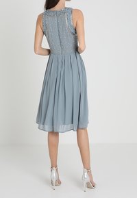 En ljusblå klänning till knäna med en plisserad kjol, pärlprydd bakparti och volang i halsringningen. Bärs med silvriga högklackade skor.