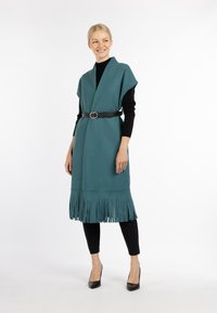 Teal fringed vest met een wikkelontwerp en V-hals, cinched in de taille met een zwarte riem. Zwarte slim-fit broek en hoge hakken maken de outfit compleet.