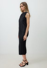Schwarzes, plissiertes Midikleid mit hohem Kragen und ärmellosem Design, das vertikale Texturen und eine figurbetonte Silhouette aufweist, kombiniert mit schwarzen, hohen Sandalen.