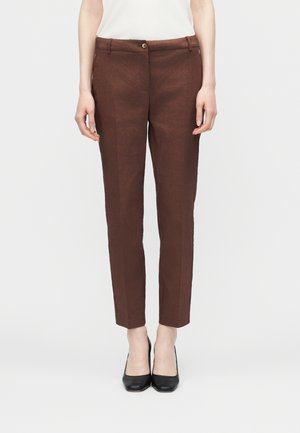 Nő vékony fazonú barna nadrágban, elöl gombbal és övbújtatóval, fekete magas sarkú cipővel és fehér felsővel, fehér háttér előtt állva.