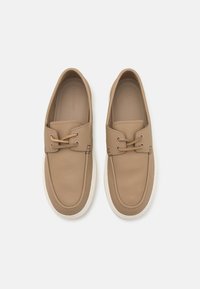 Scarpe slip-on in pelle beige con punta arrotondata, cuciture dettagliate e due fori per i lacci. Presentano una suola in gomma bianca e una parte superiore testurizzata.