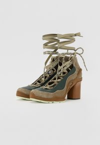 Lace-up booties in dark green and beige with a textured, sparkly finish and a chunky brown heel. Features intricate lacing and contrasting accents.