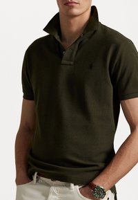 Man i en mörkgrön kortärmad polo med upprätt krage och en grön klocka, med händerna i fickorna på vita byxor.