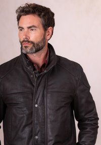 Bearded man with dark curly hair wearing a zipped black leather jacket and a red plaid shirt, looking to the left against a plain background.