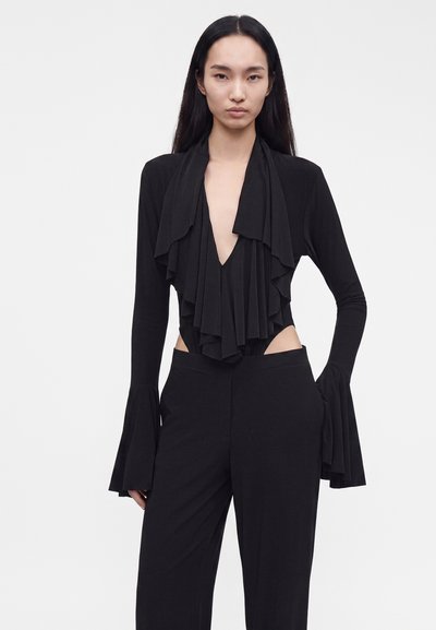 Γυναίκα με μαύρο outfit, μπλούζα με βαθύ V-λάστιχο και βολάν, μακριά φαρδιά μανίκια και μαύρο παντελόνι ψηλόμεσο σε απλό λευκό φόντο.