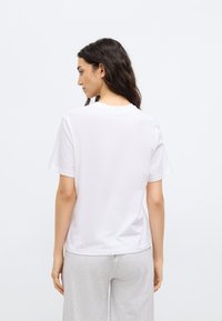 Camiseta blanca de manga corta hecha de algodón suave. Diseño sencillo con cuello redondo, textura lisa y estilo minimalista, combinada con pantalones gris claro.