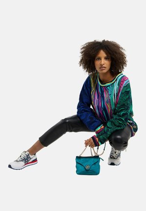 Vrouw met krullend haar die hurkt, draagt een kleurrijke gebreide trui met patroon, zwarte leren broek, witte sneakers, en houdt een turquoise handtas met gouden ketting vast.