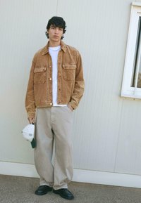Giacca in velluto a coste color ocra con chiusura a bottoni e due tasche sul petto, abbinata a pantaloni a gamba larga beige chiaro e scarpe nere. T-shirt bianca sotto.