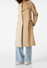 Beżowy trencz z podwójnymi guzikami, bocznymi kieszeniami i klasycznym kołnierzem. Noszony na szerokich, jasnoniebieskich dżinsach.