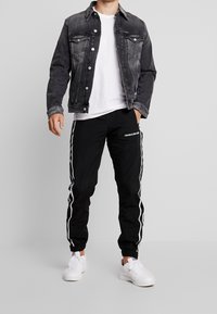 Man i svart tvättad denimjacka, vit t-shirt, svarta Calvin Klein träningsbyxor med vita sidostråk, och vita sneakers som står på grått golv.