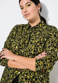 Camisa de estampado de leopardo verde con acentos amarillos, diseño abotonado y mangas enrolladas, hecha de tejido suave. El modelo cruza los brazos en pose.