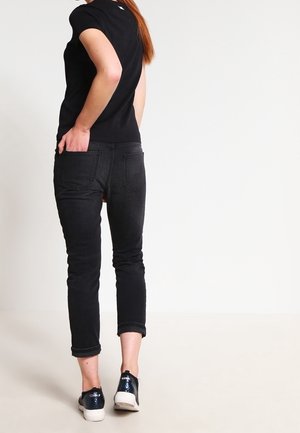 Femme portant un t-shirt noir, un jean court noir et des baskets noires avec des semelles blanches, debout devant un fond blanc uni.