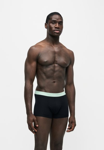 Homme musclé à la peau foncée portant un boxer noir avec une ceinture bleu clair, debout sur un fond blanc uni.