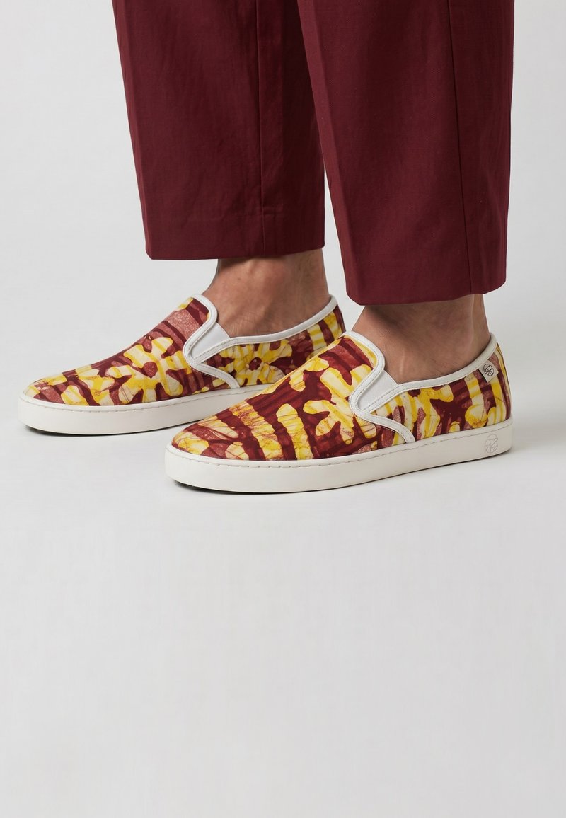 Pieds portant des chaussures à enfiler avec des motifs abstraits jaunes et rouges et des semelles blanches, associées à un pantalon court rouge foncé.
