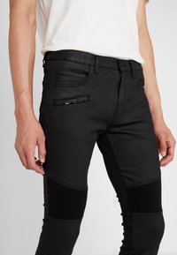 Jeans de mezclilla negra con un ajuste slim, que incluyen paneles de tela negra texturizada, bolsillos laterales y un bolsillo con cremallera en el muslo derecho.