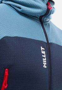 Hooded jacket in light blue and navy, featuring textured fabric, a central zipper, red zipper pull, and a white logo on the front.