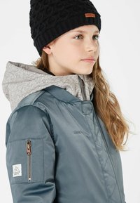 Blaue Satinjacke mit grauem Fleece-Haube, ausgestattet mit einer Reißverschlusstasche am linken Arm und einer gestrickten schwarzen Mütze.