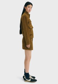Braune Cordjacke mit Seitentaschen, kombiniert mit einem passenden Cordrock. Das Modell trägt dunkle Socken und schwarze Sneakers mit weißen Akzenten.