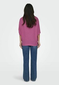 Maglione rosa fluido con maniche corte e larghe, abbinato a jeans blu a gamba dritta. Texture morbida, design casual.