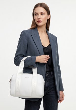 Donna che indossa un blazer grigio e un top nero tiene una borsa bianca Diana & Co con doppi manici, in piedi davanti a uno sfondo semplice.