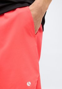 Χέρι στην τσέπη φωτεινών κοραλί σορτς με μικρό λευκό λογότυπο κοντά στο τελείωμα, φορεμένα με μαύρο τοπ.