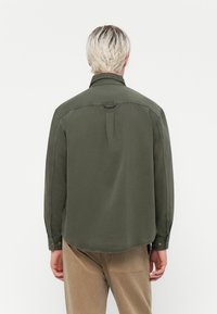 Olive green, long-sleeved shirt with a curved hem, button cuffs, and a back loop detail. Made from a smooth fabric with minimal texture.