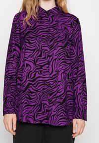 Chemise boutonnée violette avec un motif abstrait de zèbre noir. Le tissu semble lisse, avec des manches longues et un design de col classique.