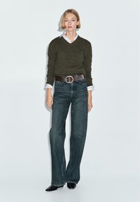 Maglione verde scuro con scollo a V, camicia bianca con colletto sotto, jeans blu a vita alta, cintura marrone con fibbia rotonda, scarpe nere.