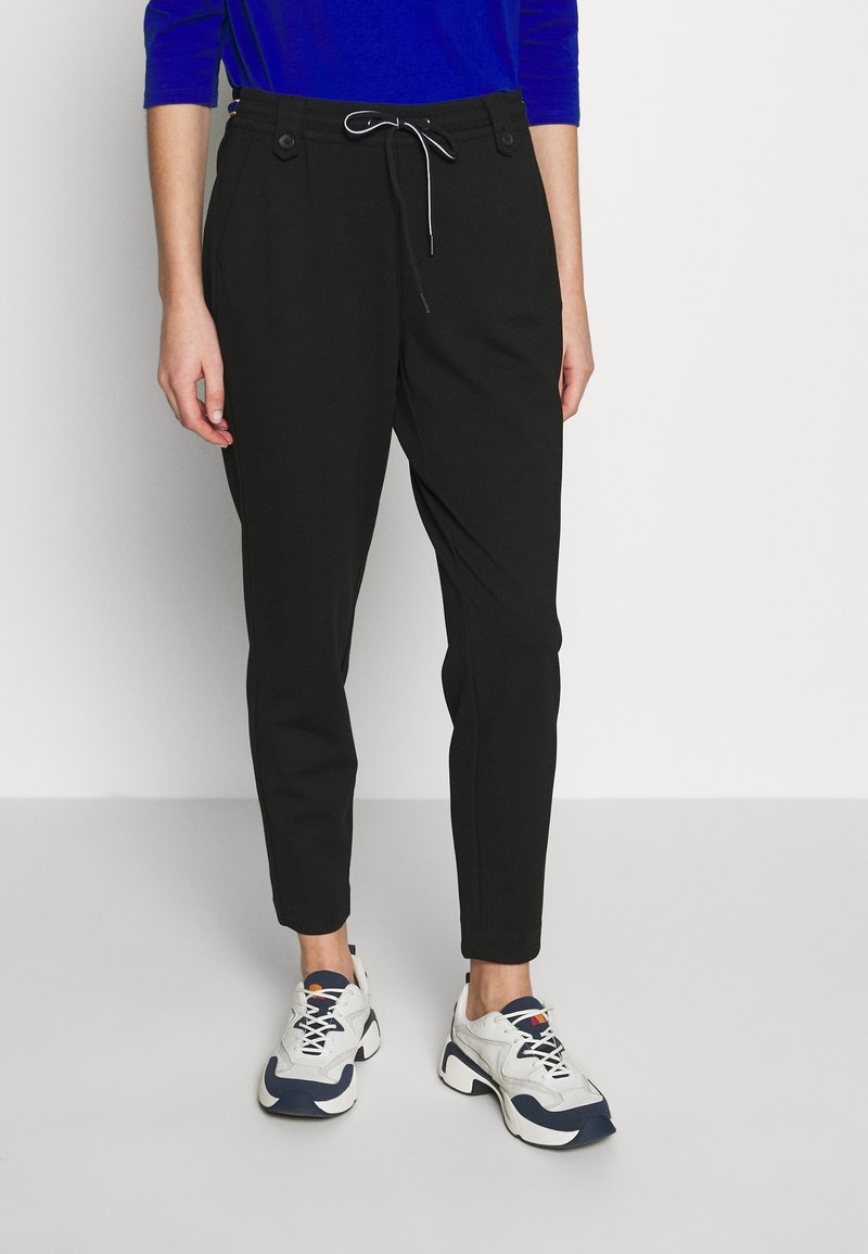 Personne portant un pantalon noir fuselé avec une taille à cordon et des baskets blanches avec des touches bleues, debout sur un fond blanc.