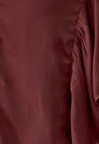 Burgundy blouse made of smooth fabric, featuring pleated accents on the sides, short sleeves, and a relaxed fit.