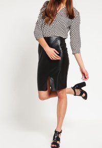 Svart faux läder blyertskjol med en framspringa, i kombination med en svart- och vitrutad blus. Svarta högklackade sandaler fulländar looken.