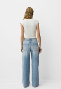 Tricou alb ajustat, cu mâneci scurte, asociat cu blugi evazați de culoare albastru deschis, cu buzunare la spate și un design cu talie înaltă. Textură netedă.