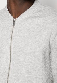 Veste aviateur grise en tissu texturé, dotée d'une fermeture éclair sur le devant et d'un col rond. Design simple sans motifs visibles.