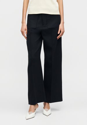 Čierne nohavice s širokými nohavicami z hladkej látky, s dvoma zadnými vreckami a zažehlenými záhybmi, doplnené krémovým topom a bielou obuvou.