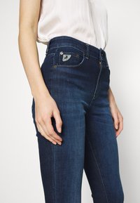 Tmavě modré denimové džíny s vysokým pasem, které mají na přední kapse malé vyšívané logo, měděné kovové detaily a úzký střih.