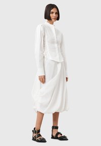 Camicia bianca a maniche lunghe con colletto alto, caratterizzata da dettagli arricciati ai lati, abbinata a una gonna bianca voluminosa e sandali neri con cinturini.