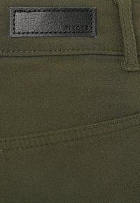 Ύφασμα ελιάς με λεία υφή· διαθέτει μια μαύρη δερμάτινη ετικέτα με την επιγραφή "PIECES" και εμφανείς ραφές.