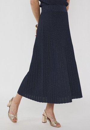 Femme portant une jupe midi plissée bleu marine et des sandales à talons carrés dorées avec brides, se tenant avec un pied légèrement levé.