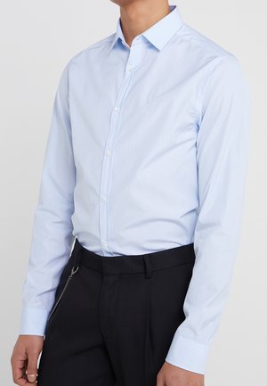 Chemise bleu clair à boutons avec un col pointu, des manches longues et un logo brodé sur la poitrine. Portée avec un pantalon noir.