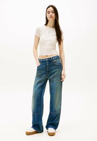 Beżowy krótki t-shirt z małym logo, w połączeniu z szerokimi niebieskimi dżinsami z przednimi kieszeniami i wyblakłym praniem. Białe trampki dopełniają całość.