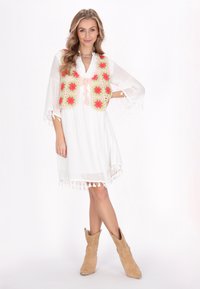 Weißes Kleid mit fransigem Saum, kombiniert mit einer bunten Häkelweste mit rosa, grünen und roten Blumenmustern. Beige Ankle Boots.